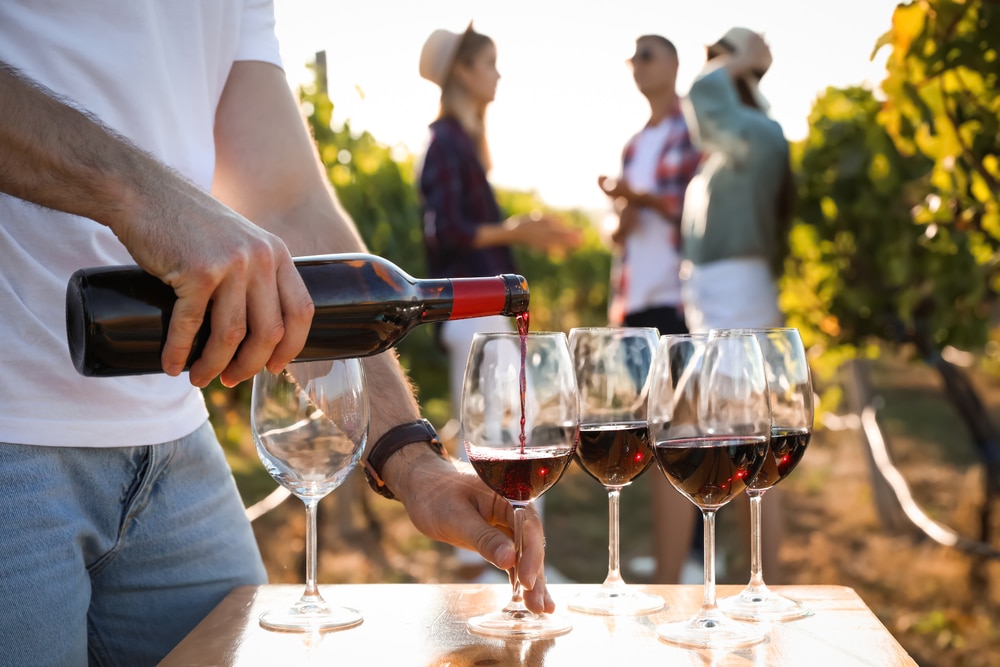 Man pouring red wine from bottle into glasses outside, friends in the background at wineries on Seneca Lake.