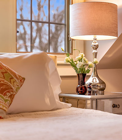 A nightstand with a lamp and vase of white flowers next to a bed, with a window in the background.