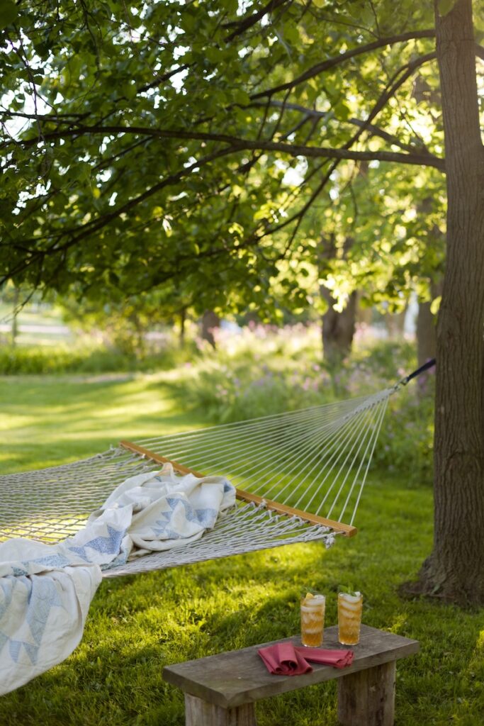 A hammock with a blanket hangs between trees in a sunny, green garden with a wooden bench holding two drinks below.
