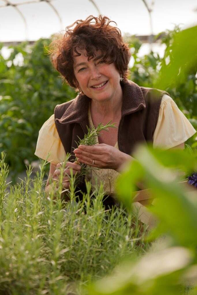 A smiling woman with curly auburn hair tends to rosemary plants in a greenhouse setting.