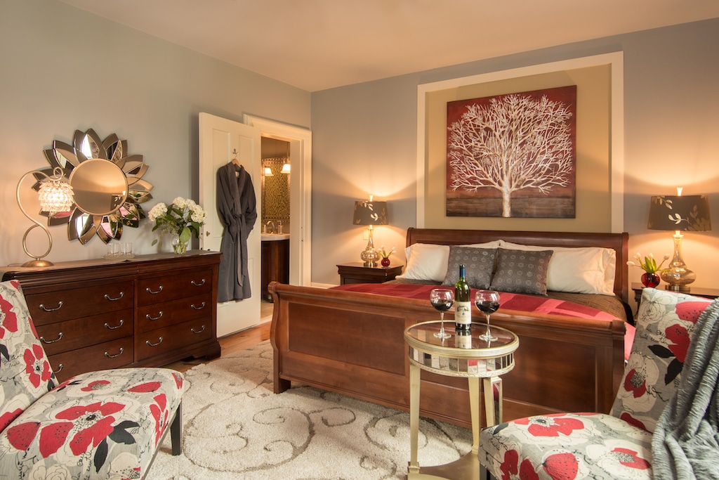 A cozy bedroom with a wooden bed, floral chairs, a dresser, and a small table with two glasses of red wine.