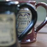Two ceramic mugs with "The Inn at Gothic Eves" logos; the front one is blurred, the back one is in focus.